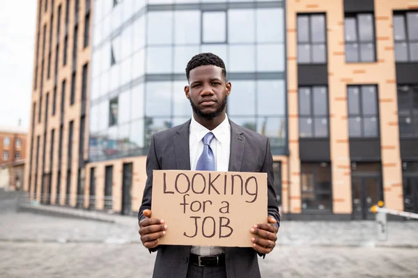 Ung svart affärsman som håller pappskylt med texten LOKING FOR A JOB, förlorade sitt jobb på grund av karantän — Stockfoto