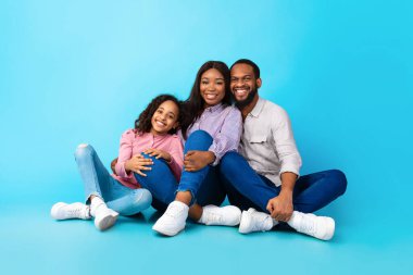 Siyah adam gülümseyen kadın ve kıza sarılıyor, yerde oturuyor.