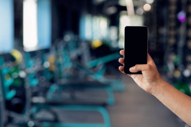 Spor salonunun arka planında boş siyah ekranlı akıllı telefon tutan erkek.