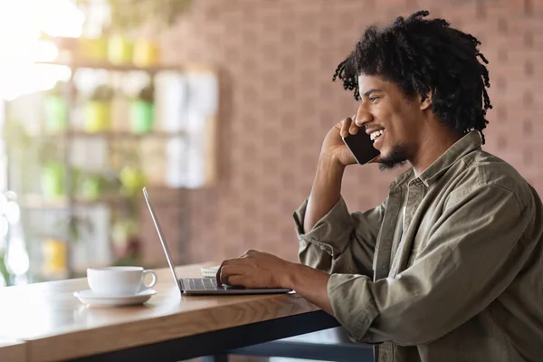 Negro freelancer chico hablando en el teléfono celular mientras trabaja con el ordenador portátil en café —  Fotos de Stock