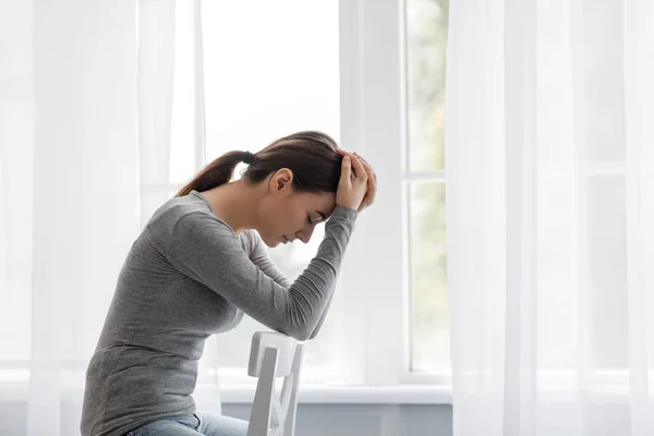 Ongelukkig europees duizendjarig vrouwtje verdrietig thuis alleen, houdt haar hoofd vast en zit op stoel — Stockfoto