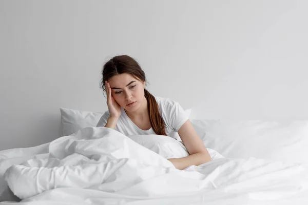 Triste mujer europea milenaria, preocupada, sufre de insomnio y dolor de cabeza. Señora se sienta en la cama en el dormitorio —  Fotos de Stock