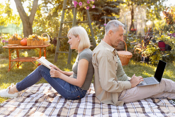 Ушедшая в отставку супружеская пара отдыхает в саду, женщина читает книгу, в то время как мужчина использует ноутбук, сидя спиной к спине, вид сбоку