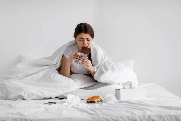 Smutny głodny biały młoda dama jedzenie czekolada bar na łóżku z fast food w domu, cierpiących na stres — Zdjęcie stockowe