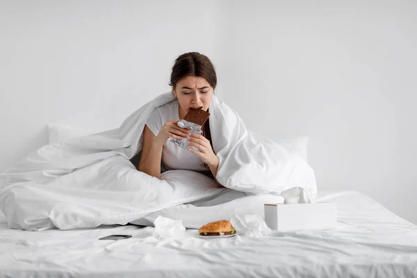 Sad hungry caucasian young lady eating chocolate bar on bed with fast food at home, suffering from stress