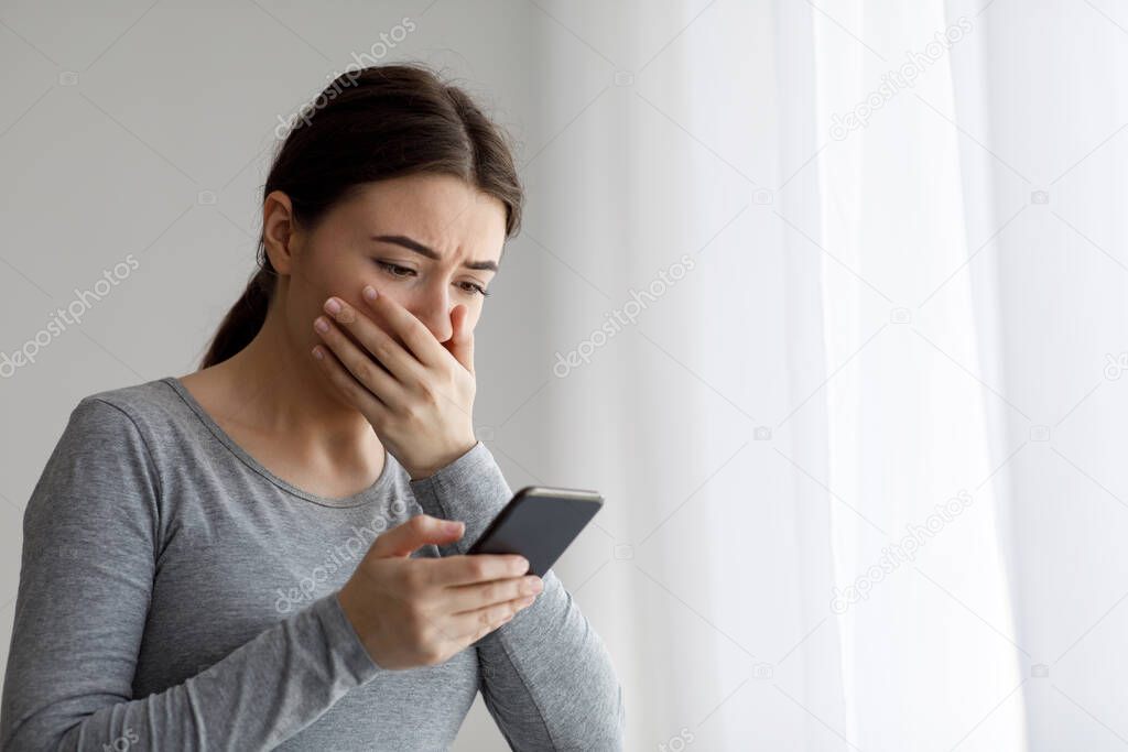 Upset depressed millennial pretty woman covering her mouth with hand and looking at phone