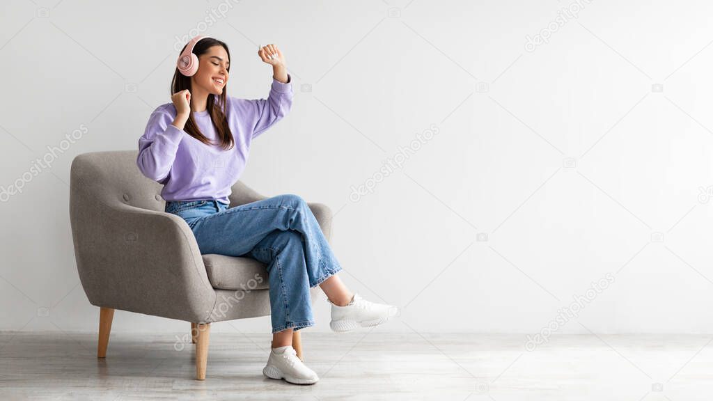 Cheery young lady in headphones listeing to music and dancing while sitting in armchair against white studio wall