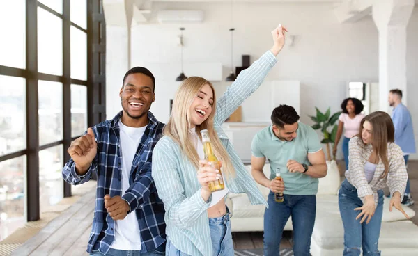 Cheery blonde lady singing and dancing, using bottle of beer as microphone, partying with her diverse friends at home — Stock Photo, Image