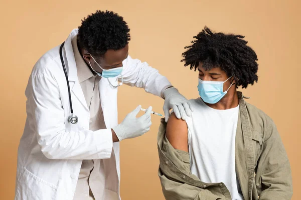 Black Doctor Making Vaccine Shot To Male Patient Wearing Medical Mask