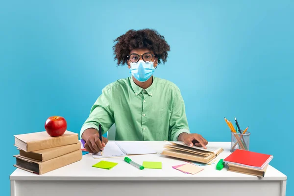 Education during pandemic. African american male student in protective face mask sitting at table and doing homework