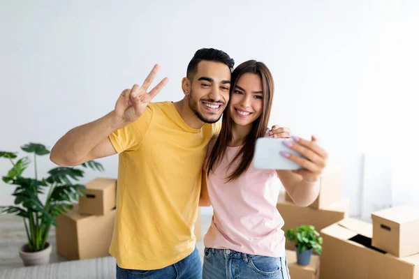 Sonriente chico árabe y su novia caucásica mudándose a su propia casa juntos, tomando selfie entre cajas de cartón — Foto de Stock