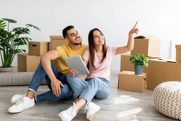 Feliz pareja interracial con tableta PC sentado en el suelo entre cajas de cartón y el diseño de planificación de su nuevo apartamento —  Fotos de Stock