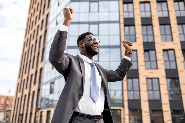 Spännande afrikansk amerikansk affärsman firar framgång och räcker upp händerna, står utomhus nära kontorshotell — Stockfoto