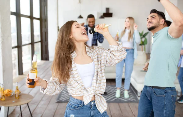 Młoda blondynka imprezuje ze swoimi różnorodnymi przyjaciółmi, śpiewa karaoke do mikrofonu, tańczy i pije piwo w domu — Zdjęcie stockowe