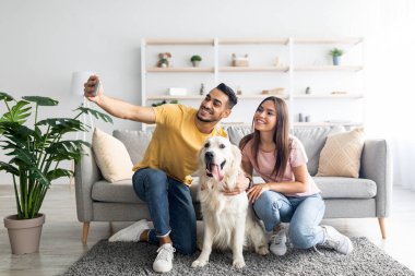 Mutlu ırklar arası çiftler köpekleriyle selfie çekerken evlerindeki yumuşak halıda oturuyorlar.