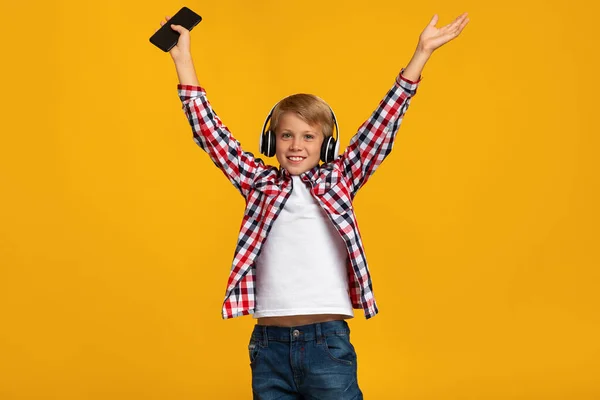 Glad for at tenåringsgutt med hodetelefoner holder smarttelefon, hopper, danser og løfter opp hendene – stockfoto