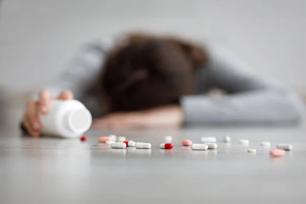 Неузнаваемая молодая белая женщина лежит на полу с банкой и разбросанными таблетками на полу, совершая самоубийство — стоковое фото