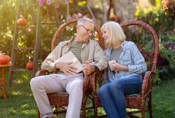 Loving elderly spouses resting in garden and enjoying warm autumn days, sitting in wicker chairs outdoors, copy space — Stock Photo, Image