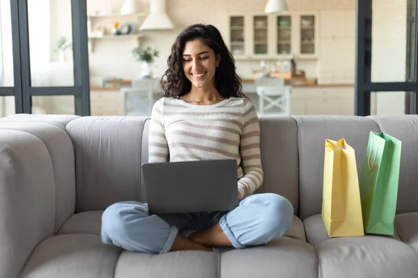 Compras en línea. Mujer joven con ordenador portátil y bolsas de compras que ordenan bienes en la web mientras está sentado en el sofá en casa — Foto de Stock