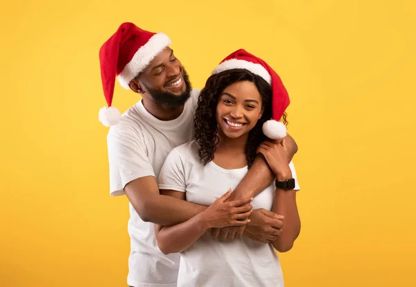 Loving african american couple in Santa hats hugging on yellow studio background, celebrate New Year or Christmas — Stok Foto