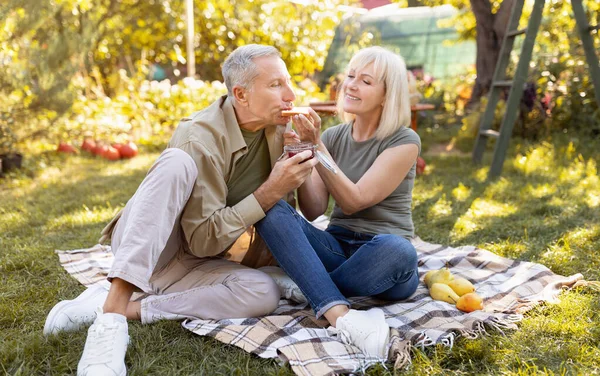Romantiskt äldre par som har picknick i trädgården, sitter på filt och äter rostat bröd, tillbringar tid utomhus — Stockfoto