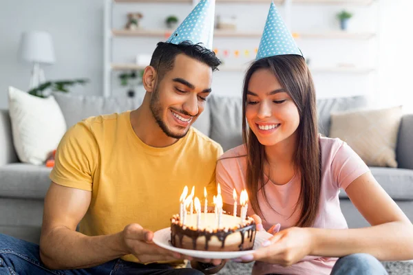 Portret van gelukkig jong multiraciaal paar in feestelijke hoeden met verjaardagstaart met aangestoken kaarsen thuis — Stockfoto