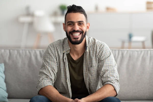 Retrato del joven árabe guapo sonriendo y mirando a la cámara, sentado en el sofá en casa — Foto de Stock