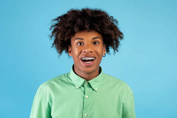 Excité afro-américain adolescent guy avec accolades regarder et sourire à la caméra, posant sur fond bleu studio — Photo