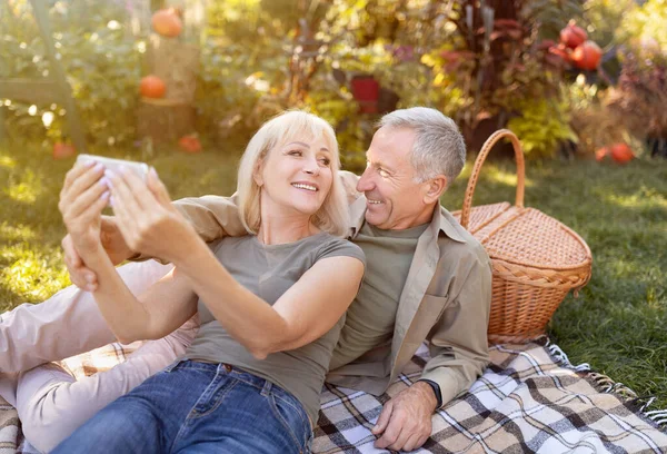 Modern elderly spouses taking selfie on smartphone, having picnic and resting in garden at sunny autumn day