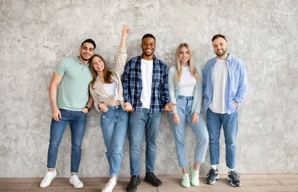 Portrait complet de jeunes amis multiraciaux posant et souriant à la caméra, debout contre le mur gris du studio — Photo