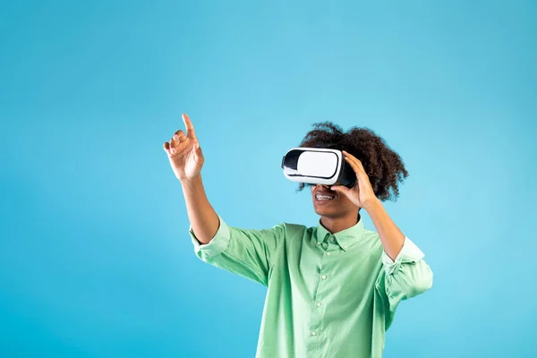 Virtual world in 3D simulation. Black teen guy in vr glasses playing and trying to touch something over blue background — Stock Photo, Image