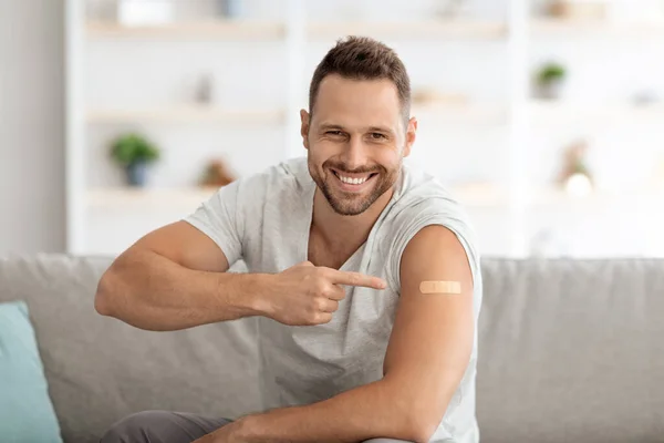 Jonge gevaccineerde man die naar de schouder wijst met medisch gips en breed glimlacht, zich gezond voelt na vaccinatie — Stockfoto