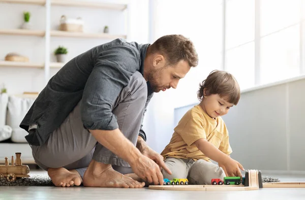 Οικογενειακό παιχνίδι. Ευτυχισμένος ενδιαφερόμενος πατέρας παίζει με το μικρό γιο, ιππασία ξύλινο τρενάκι στο σιδηρόδρομο, κάθεται στο πάτωμα — Φωτογραφία Αρχείου