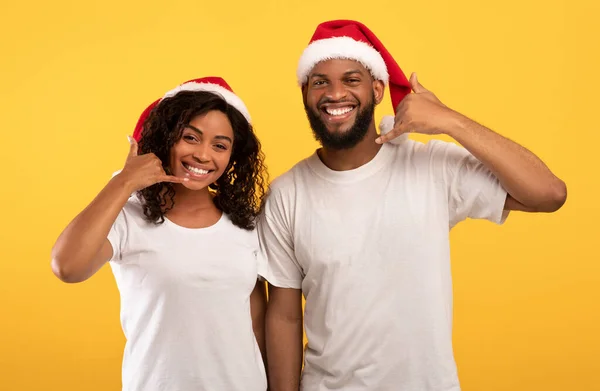 Emocionada joven pareja negra en los sombreros de Santa mostrando LLAME gesto, posando y sonriendo a la cámara en el fondo amarillo — Foto de Stock