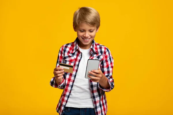 Alegre adolescente caucásico con tarjeta de crédito y teléfono de pago y compra, aislado sobre fondo amarillo — Foto de Stock