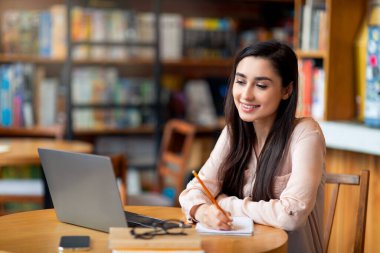 Uzaktan eğitim. Laptop kullanan ve notlar alan mutlu Latin kız öğrenci, kafenin içinde oturuyor, kopyalama alanı