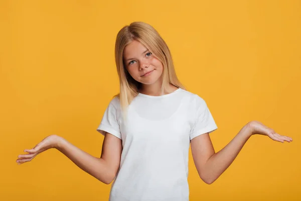 Calm confused pretty european teen girl in white t-shirt spreads her arms to the sides, isolated on yellow background — Stock Photo, Image