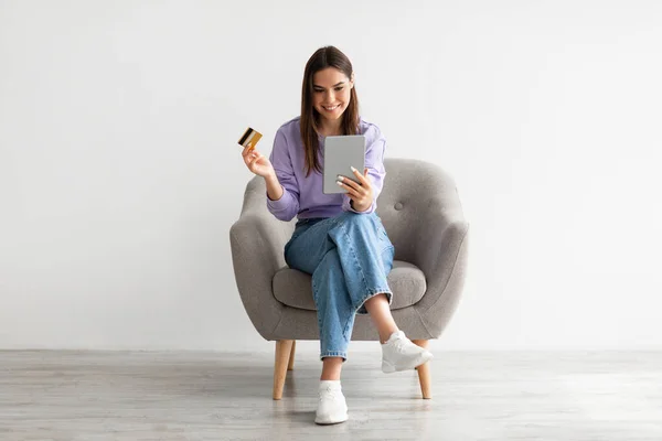 Cool joven dama con tableta digital y tarjeta de crédito de compras en línea, sentado en sillón contra la pared del estudio blanco —  Fotos de Stock