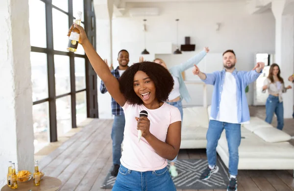 Millennial černošky dáma s lahví piva a mikrofon zpěv karaoke s různými přáteli doma — Stock fotografie