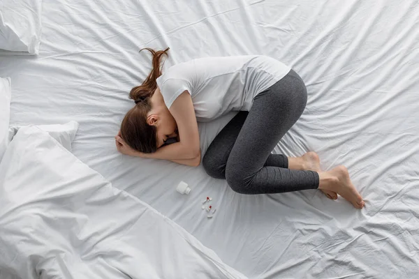 Smutna rozpacz młodej białej kobiety cierpiącej na ból brzucha leżącej na łóżku ze słoikiem i rozrzuconymi tabletkami — Zdjęcie stockowe