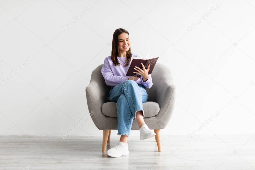 Weekend pastimes. Young Caucasian woman with open book sitting in comfy armchair against white studio wall