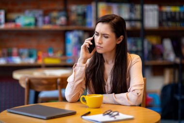 Sinirli serbest çalışan Arap bayan, cep telefonuyla müşterilerle konuşuyor, kafede uzaktan çalışıyor, dizüstü bilgisayar kullanıyor.