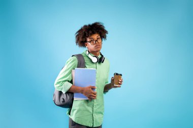 Not defterleri, paket kahve ve sırt çantasıyla yorgun erkek öğrenci mavi stüdyo arka planındaki kameraya bakıyor.