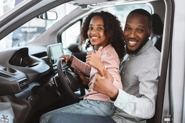 Positivo hombre negro y su linda hija sentado en el asiento del conductor del coche nuevo, mostrando pulgares hacia arriba en concesionario de automóviles —  Fotos de Stock