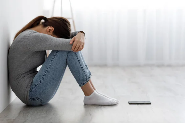 Smutná, nešťastně deprimovaná, tisíciletá běloška sedící na podlaze s telefonem a zakrývající si tvář — Stock fotografie