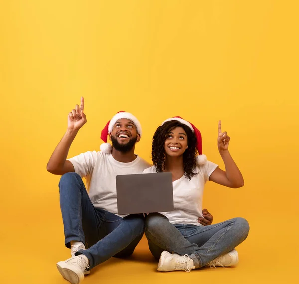 Positiva afrikanska amerikanska makar i Santa hattar sitter med bärbar dator och pekar upp på tomt utrymme på gul bakgrund — Stockfoto