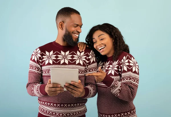 Pasangan kulit hitam yang bersemangat dengan sweater hangat menggunakan tablet digital di atas latar belakang biru, melakukan belanja Natal daring — Stok Foto