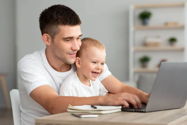 Apasági szabadság. Fiatal férfi dolgozik a laptopon otthon a csecsemő fia — Stock Fotó