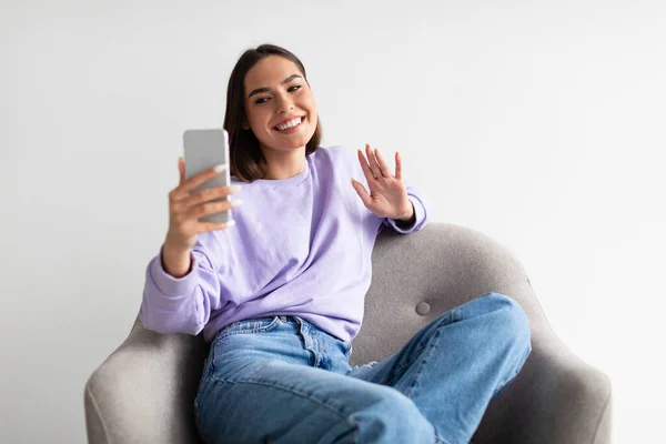 Mujer joven sonriente usando teléfono inteligente en el sillón, saludando a la webcam, hablando con amigos o suscriptores en línea — Foto de Stock