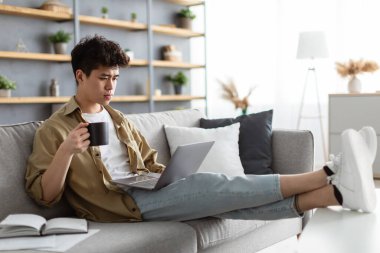 Ciddi Asyalı adam bilgisayardan video izliyor, sıcak kahve içiyor.